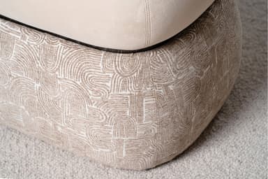 A two-tiered ottoman with a rounded, tufted top cushion and a patterned base. The top cushion is upholstered in a smooth, light beige fabric and features a single button tuft in the center, outlined by stitched seams. A contrasting piping trim defines the edge of the top cushion. The base is wider than the top and is upholstered in a textured fabric with a repeating abstract pattern in shades of beige and white, resembling arcs and linear shapes. The ottoman is situated on a textured, light-colored carpet. — 3D model