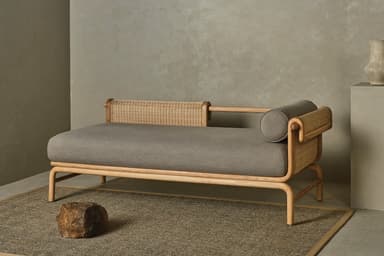 A contemporary daybed featuring a light-toned wooden frame, woven rattan detailing on one side, and a thick gray upholstered mattress with a matching cylindrical bolster pillow. The frame has gently curved edges, and the legs are integrated into the overall structure. — 3D model
