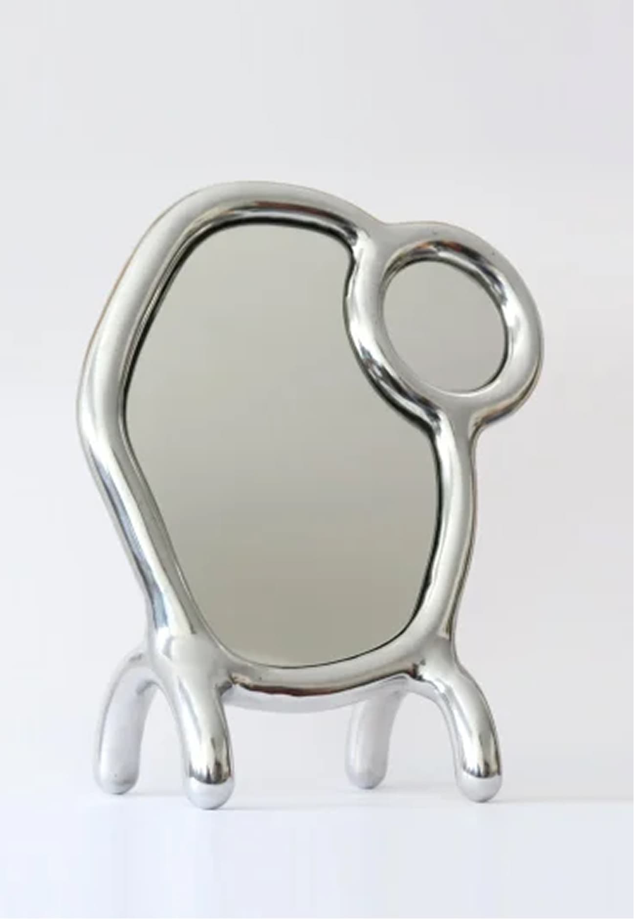 This is a unique, sculptural table mirror with an avant-garde design. It features a large, irregularly shaped reflective surface complemented by a smaller circular mirror embedded within its frame. The entire piece is supported by four bulbous, organic-shaped legs, giving it a whimsical and modern aesthetic. The highly polished metallic finish enhances its contemporary appeal and makes it a striking decorative object. — 3D model