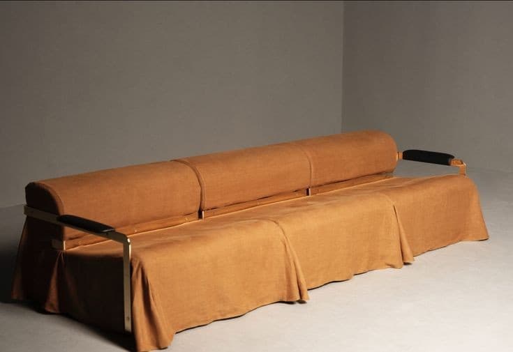A long, rectangular sofa with a curved bolster backrest. The sofa is upholstered in a textured, light brown fabric. It features a metal frame with armrests that have black accents. The base of the sofa is covered by a draped skirt of the same fabric, creating a segmented appearance. — 3D model