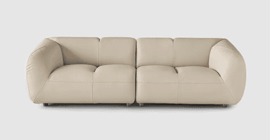 A contemporary four-seat sofa with a low profile and rounded arms. The sofa is upholstered in a light beige or cream-colored material, likely leather or a high-quality faux leather, and features prominent stitching that delineates the seat and back cushions. The design is modular, with two distinct seating sections joined together. It is supported by small, dark, cylindrical feet. — 3D model