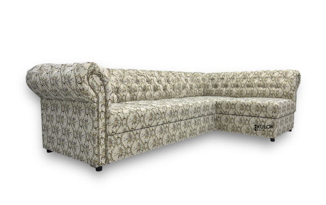 A sectional sofa with a distinctive tufted backrest and rolled arms. The sofa is upholstered in a cream-colored fabric featuring a detailed floral pattern in muted tones of green, yellow, and purple. It has a low profile with dark, block-style feet. The design suggests a Chesterfield influence with its deep buttoning and tailored appearance. — 3D model