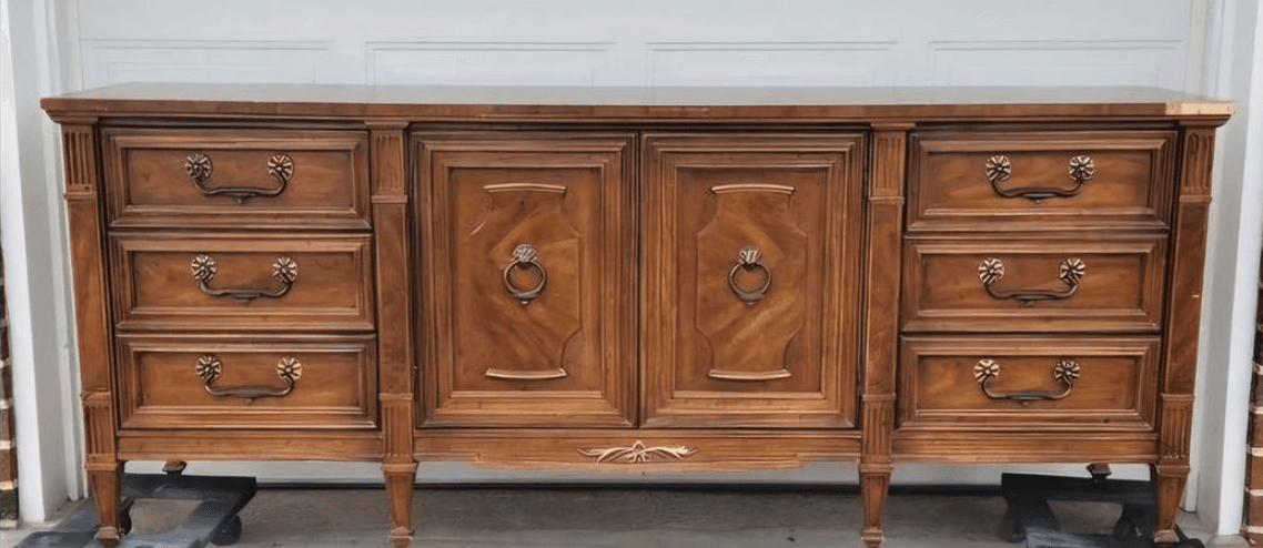 A long, wooden dresser with a traditional design. It features two cabinet doors in the center, each with a ring pull handle and ornate detailing. On either side of the doors are stacks of drawers, with three drawers visible on the left and three on the right. The drawers have decorative pulls and recessed panels. The dresser has fluted columns on the sides and rests on short, tapered legs. There is a decorative brass accent piece on the lower front panel. — 3D model