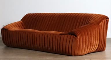 A plush, channel-tufted sofa upholstered entirely in a warm, burnt orange velvet fabric. It features a low-profile design with rounded contours and integrated armrests that seamlessly flow into the backrest and base. The distinctive vertical channel tufting provides a rich texture across all upholstered surfaces. — 3D model