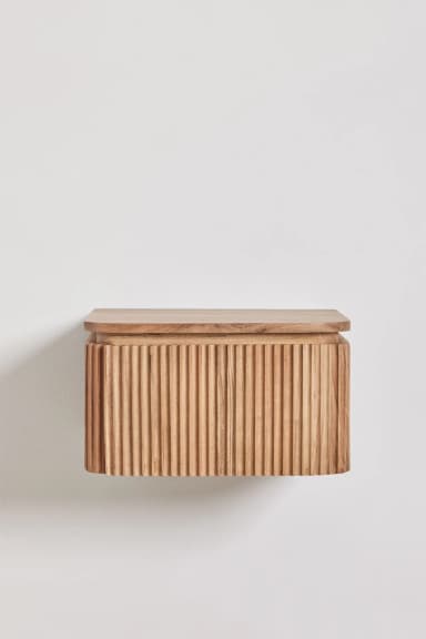A wall-mounted rectangular storage unit made from natural light-toned wood. The front and sides feature a distinctive vertical fluted or reeded texture, while the top surface is smooth with gently rounded edges, creating a soft, contemporary aesthetic. It appears to function as a single drawer or a door, though the opening mechanism is not explicitly visible. The unit is designed to float, attaching directly to a wall, offering a minimalist profile and saving floor space. — 3D model