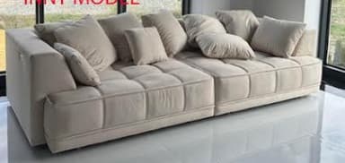 A modular sectional sofa with a low profile. It features a light-colored fabric upholstery with a tufted or channel-stitched pattern on the seat cushions and the base. Multiple throw pillows in a matching fabric are arranged on the backrest, some are square and others are slightly more rectangular. The sofa appears to be composed of at least two connected sections, forming an L-shape or a long linear arrangement. The design is contemporary and minimalist. — 3D model