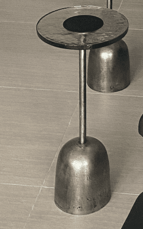 A small, industrial-style metal table featuring a round top with a raised rim and a central dark insert, supported by a cylindrical stem that widens into a bell-shaped, solid base. The metal has a textured, dark gray finish. — 3D model