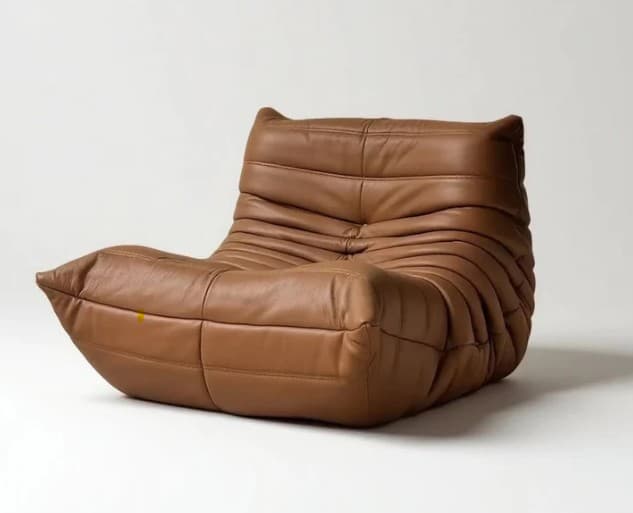 A low-profile, single-seat armchair with a distinctive rounded, organic form. The chair is upholstered in brown leather with deep channel tufting that creates a visually textured and plush appearance. The armrests and seat appear to be a continuous, flowing element, with no distinct legs visible, suggesting it rests directly on the floor or a low base. — 3D model