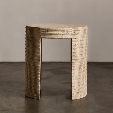A cylindrical end table with a textured, stone-like surface. The table has a rounded top and a single, curved leg or base element visible. The side of the table exhibits a series of horizontal grooves or ridges, suggesting a layered construction or decorative carving. The material appears porous and natural, consistent with travertine or a similar stone composite. The color is a light, earthy beige. — 3D model