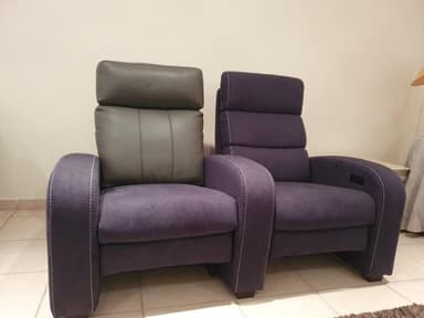 A pair of connected recliners upholstered in a tan leather-like material. Each recliner features a high back with segmented cushioning and padded armrests. The recliner on the right shows a visible control panel embedded in the armrest, suggesting electronic reclining functionality. The overall design is modern and appears to be intended for home theater or lounge seating. — 3D model