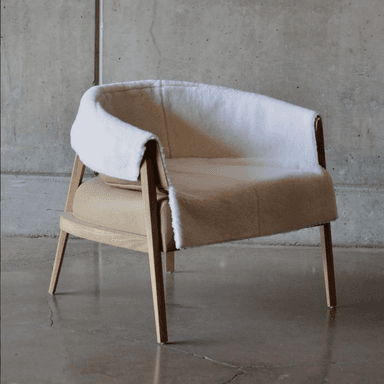 A contemporary armchair with a distinctive silhouette. The frame is constructed from light-colored wood, featuring four tapered legs and an open arm structure. The seat and back are upholstered in what appears to be tan leather, complemented by a plush, off-white sheepskin or faux shearling cover that drapes over the back and arms, creating a cozy aesthetic. The design suggests a blend of minimalist structure with textural richness. — 3D model