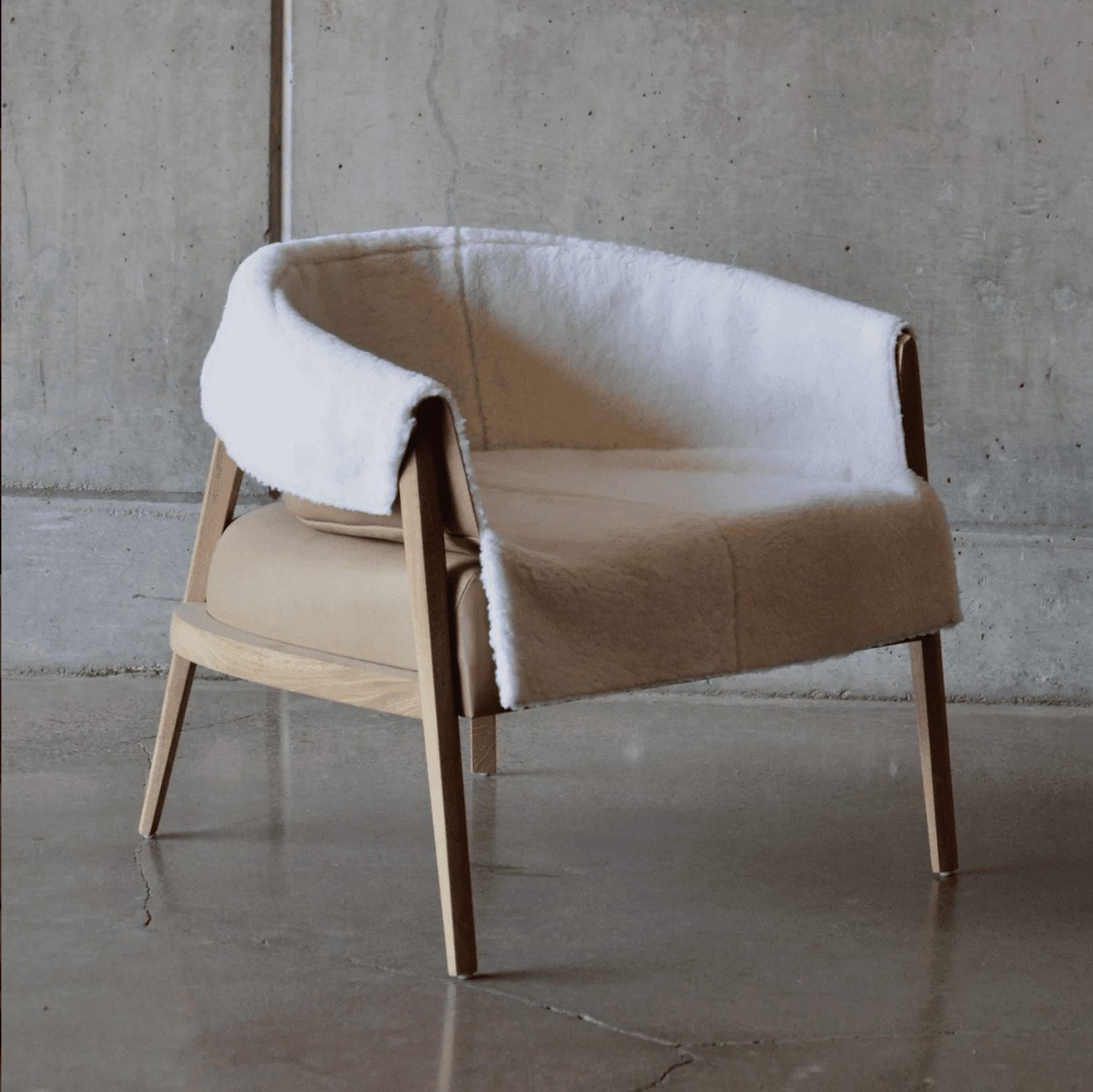 A contemporary armchair with a distinctive silhouette. The frame is constructed from light-colored wood, featuring four tapered legs and an open arm structure. The seat and back are upholstered in what appears to be tan leather, complemented by a plush, off-white sheepskin or faux shearling cover that drapes over the back and arms, creating a cozy aesthetic. The design suggests a blend of minimalist structure with textural richness. — 3D model