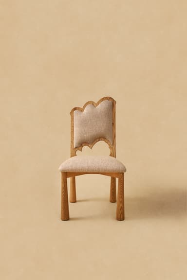 A modern armless chair featuring a wooden frame and upholstered seat and backrest. The chair's legs are stout and cylindrical, tapering slightly towards the floor. The backrest is gently curved, and both the seat and back are covered in a light beige, textured fabric. The wood has a natural, light tone, possibly oak or ash, with visible grain. — 3D model