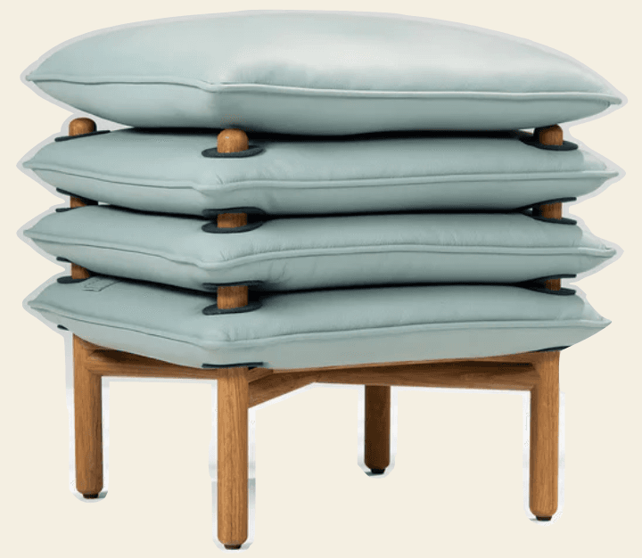 A modern ottoman featuring a stacked cushion design atop a wooden base. The cushions appear to be upholstered in a light blue leather or similar material, with subtle stitching detail. They are supported by wooden dowels and accented with dark circular elements. The base consists of four angled wooden legs connected by a frame. — 3D model