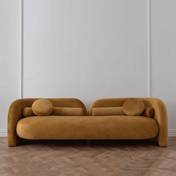 An elegant, contemporary armless sofa with a distinct curved silhouette, upholstered in a soft, plush ochre velvet fabric. It features a continuous, undulating backrest and arm-like extensions that gracefully curve around the seating area. The sofa is accompanied by one cylindrical bolster pillow and one circular pillow, designed to complement its organic form and enhance comfort. The design emphasizes fluidity and softness, suitable for modern living spaces. — 3D model