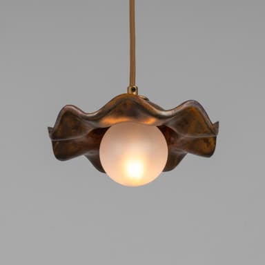 A pendant light featuring a distinctive wavy, shell-like metal shade with a mottled, patinated bronze finish. Suspended beneath the shade is a spherical frosted glass diffuser, emitting a soft, warm light. The fixture hangs from a braided textile cord, connecting to the shade via a small brass fitting. — 3D model
