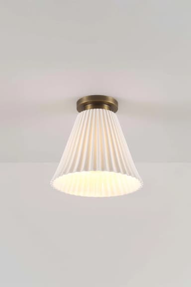 A white pleated bell-shaped light fixture mounted flush to a white ceiling. The fixture features a matte brass circular plate that attaches to the ceiling, from which the fluted, conical shade extends downwards. The light source within the shade is illuminated, casting a warm glow and highlighting the vertical pleats. — 3D model