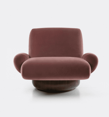 A plush armchair with a rounded silhouette and voluminous cushioning. The seat and backrest are upholstered in a soft, rose-colored velvet fabric. It features integrated armrests that flow seamlessly from the main body of the chair. The chair is supported by a substantial, dark-stained wooden base with a rounded, drum-like shape. — 3D model