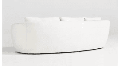 A close-up view of a cream-colored sofa, focusing on the armrest and seat cushions. The upholstery appears to be a textured boucle or similar woven fabric. The armrest has a rounded, curved profile, and the seat cushions are plush with visible stitching. The base of the sofa, where it meets the floor, is partially visible and appears to be a dark wood. — 3D model