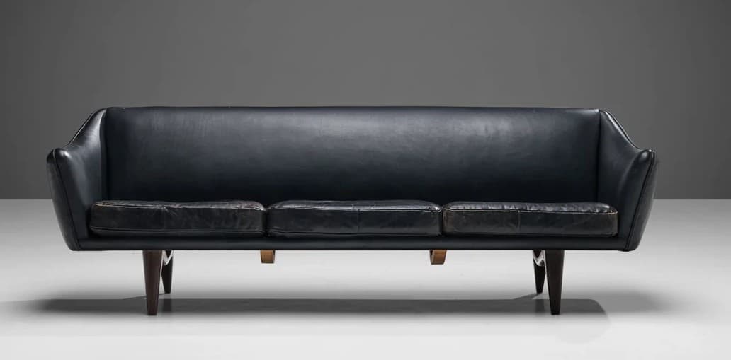 A three-seater sofa with a curved backrest and armrests, upholstered in dark leather. The seat is composed of three distinct cushions. The sofa is supported by four tapered wooden legs. — 3D model