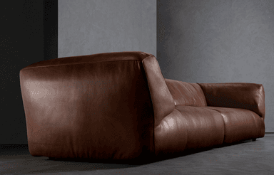 A low-profile, two-seater sofa upholstered in brown leather. The sofa features a rounded, pillow-like design with plush cushions for the seat and back. The arms are integrated into the body of the sofa and curve outwards. The base appears to be a dark, slim frame. — 3D model