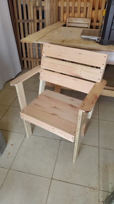 An unfinished slatted wooden armchair, constructed from light-colored timber boards. The design features a straight back, integrated armrests, and a flat seat, all composed of horizontal wooden slats. The visible fasteners suggest a simple, utilitarian construction. — 3D model