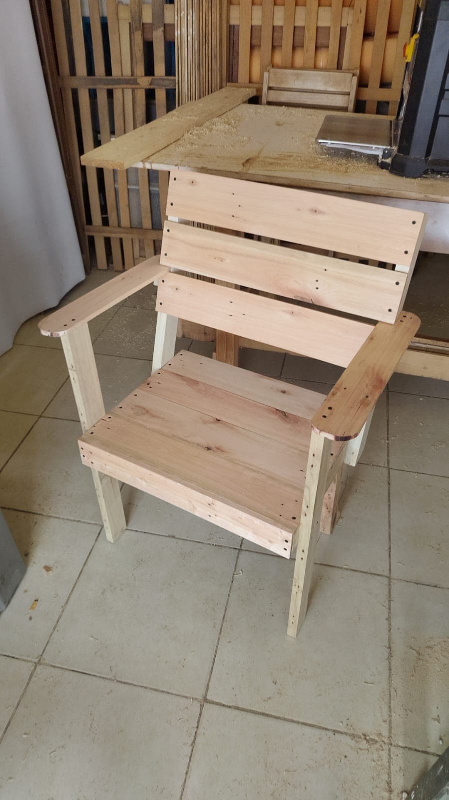 An unfinished slatted wooden armchair, constructed from light-colored timber boards. The design features a straight back, integrated armrests, and a flat seat, all composed of horizontal wooden slats. The visible fasteners suggest a simple, utilitarian construction. — 3D model