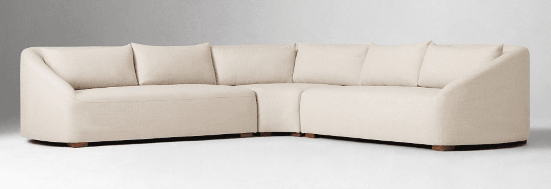 A sectional sofa composed of multiple modules, featuring a cream-colored linen-like upholstery. The sofa has rounded arms and a curved back on one end. It is supported by short, dark wooden block feet. The seating area is furnished with several plush back cushions. — 3D model