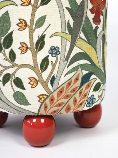 A compact, cylindrical ottoman upholstered in a multi-colored fabric featuring an intricate Arts and Crafts-inspired floral and botanical pattern. The main body of the ottoman rests on four short, spherical, brightly lacquered red feet, providing a contrasting base. The fabric design includes motifs of red flowers, green leaves, and delicate tendrils on an off-white background. — 3D model