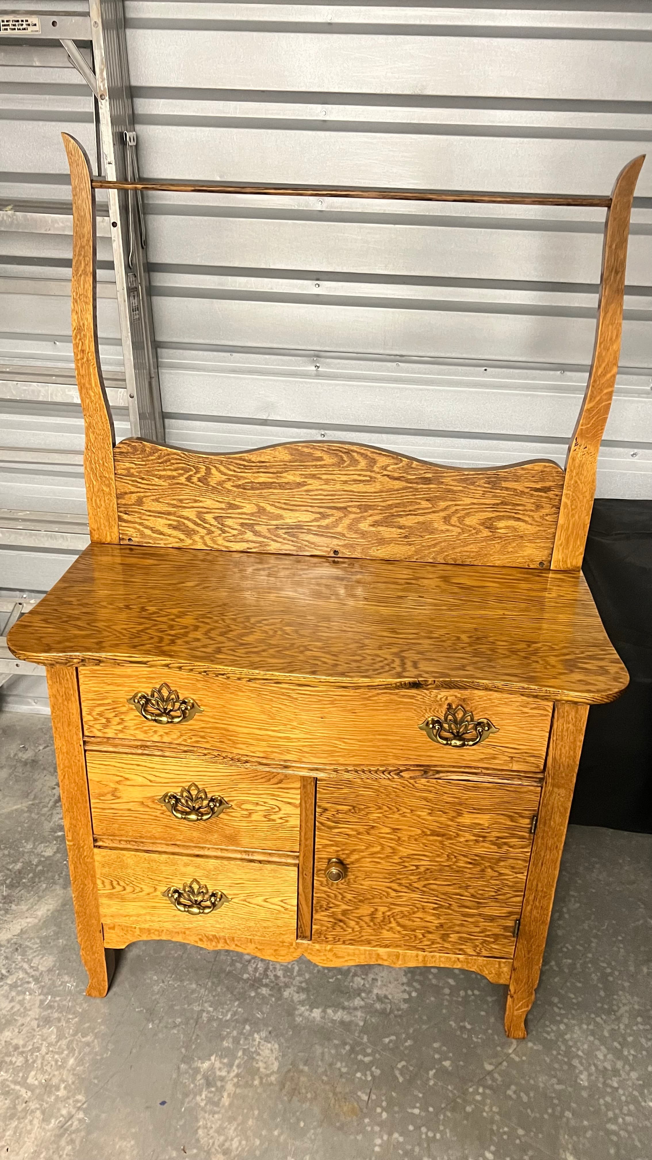 A traditional golden oak washstand featuring a combination of storage with two drawers on the left and a single cabinet door on the right, all adorned with ornate brass pull handles. The top surface is flat and polished, and an elevated back panel with a scalloped top edge supports a horizontal wooden towel bar. The piece stands on four shaped legs, with the front two exhibiting a gentle curve. The wood grain is prominent, indicative of solid oak construction. — 3D model