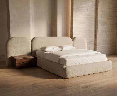 A contemporary upholstered bed featuring a low-profile frame and an extended, multi-panel headboard with rounded edges. A dark wood nightstand is positioned next to the bed, appearing to be integrated into the overall design. The bed and headboard are covered in a light beige, textured fabric. — 3D model