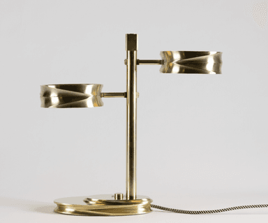 A modern two-light table lamp constructed from brass, featuring a central vertical stem with two horizontally extending arms, each supporting a cylindrical light fixture with a distinctive fluted or reeded exterior texture. The base is an oval, layered design with similar fluted detailing. The lamp is illuminated, showcasing integrated LED light sources, and includes a braided fabric power cord. — 3D model