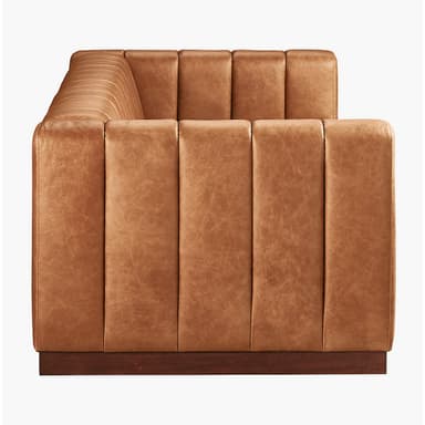 A long, channel-tufted sofa with a minimalist design. The sofa features a continuous padded back and armrest with vertical channels, upholstered in a tan leather-like material. It rests on a low, dark wood plinth base. The overall aesthetic is modern with a touch of art deco. — 3D model