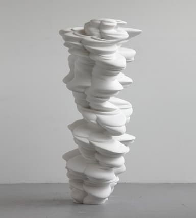 A tall, abstract floor sculpture with an organic, twisted form, rendered in a smooth, polished white material, likely resin or fiberglass. It stands on a simple square base, illuminated by uplighting, within an interior space characterized by ancient-looking brick walls. — 3D model