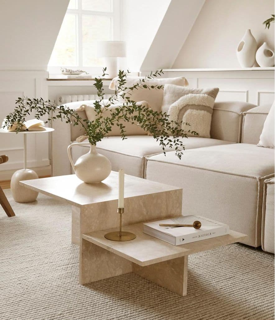 A geometric, tiered coffee table with intersecting planes, appearing to be constructed from a light-colored stone or a similar composite material. It features two main horizontal surfaces at different heights, supported by a cross-base structure. A cream-colored vase with green foliage, a slender white candle in a brass holder, and a small book with a wooden spoon are placed on its surfaces, suggesting its use as a decorative and functional piece in a living space. — 3D model