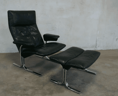 A black leather armchair with a matching ottoman. The chair features a high back and a curved, cantilevered chrome base. The ottoman also has a curved chrome base and is upholstered in matching black leather with a segmented cushion design. — 3D model