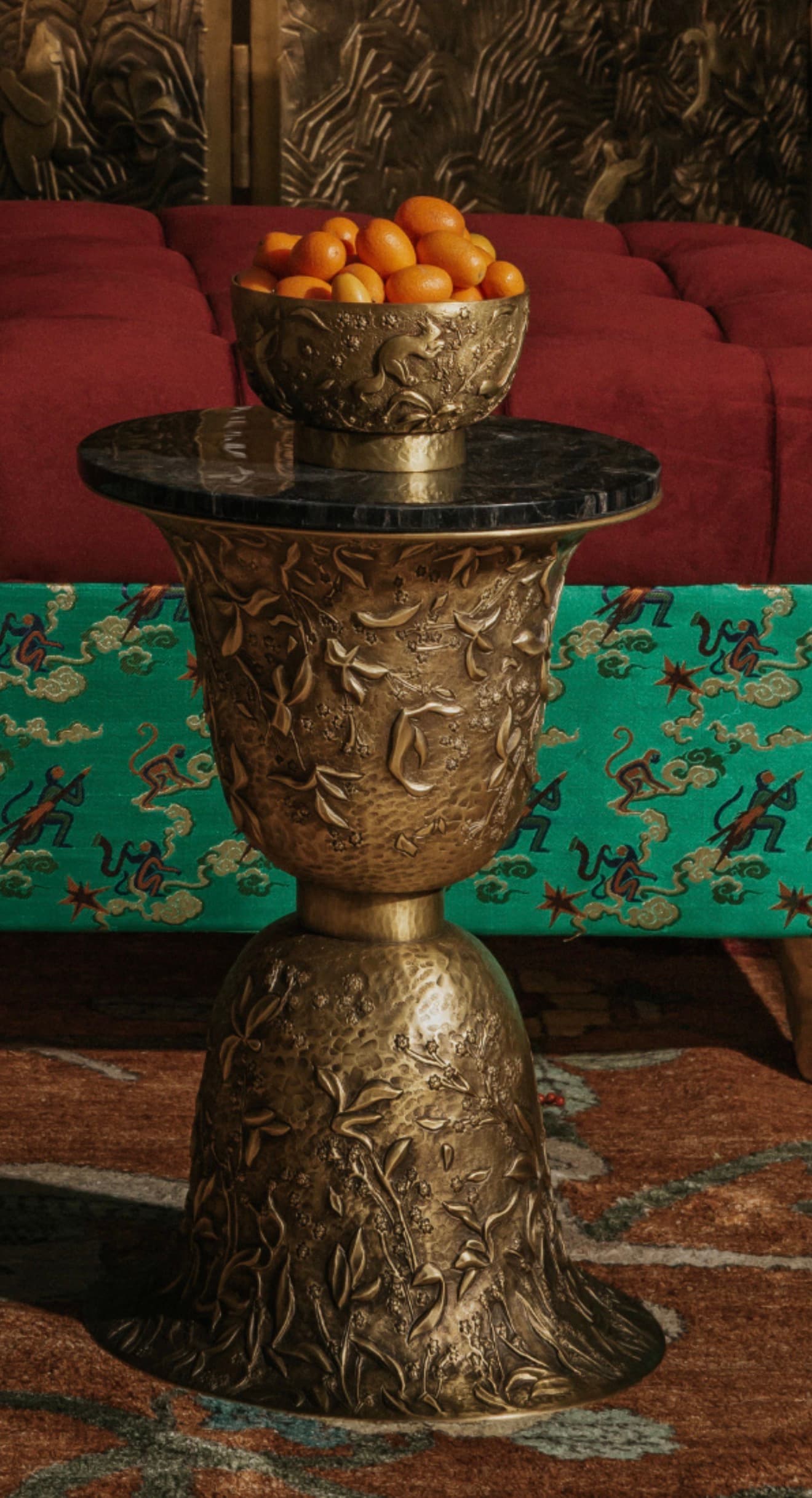A highly ornate pedestal-style side table featuring a dark, polished black marble tabletop. The base is crafted from a golden-toned metal, possibly brass or bronze, and is extensively decorated with deeply embossed botanical motifs and a distinctive hammered texture. The table stands on a flared, bell-shaped base, with a central column supporting the circular marble top. A smaller, also ornate, metal bowl filled with kumquats rests on the tabletop. — 3D model