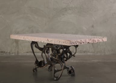 A console table with a rectangular, light pinkish-beige stone top and an intricately sculpted, dark bronze base. The base features an organic, abstract design with interconnected hollow forms and flowing lines, resembling bones or skeletal structures. The stone top has a rough-hewn edge and a textured surface with visible inclusions. — 3D model