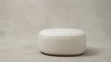 A contemporary upholstered ottoman with a rounded oval shape, covered in a plush cream bouclé fabric. The design features soft, curvilinear forms and a low-profile base, contributing to its modern and inviting aesthetic. A subtle seam runs along the perimeter of the top surface, connecting to vertical seams on the sides. — 3D model