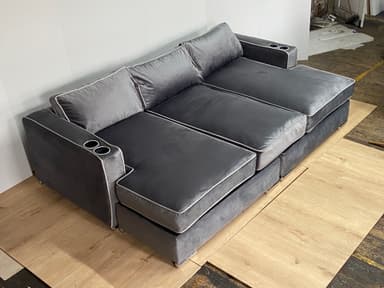 A modern sectional sofa with a chaise lounge extension on one side. The sofa features multiple seat cushions and back cushions, upholstered in a soft, plush gray fabric. It has a piped trim in a lighter contrasting color along the edges of the cushions and frame. Two integrated cup holders are visible on the armrests. The base appears to be a solid structure, possibly with decorative metal feet. — 3D model