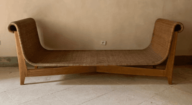 A contemporary chaise lounge with a sinuous, ergonomic design. The frame is constructed from light-colored wood, featuring gently curved legs and supports. The seating surface and backrest are made of a woven material, possibly rattan or cane, creating a textured appearance. The headrest area is formed by a continuous, rolled curve of the woven material. — 3D model