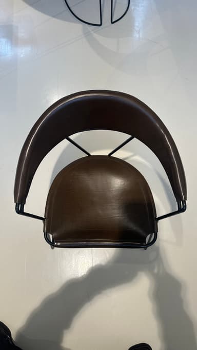 A contemporary armchair with a dark brown leather seat and backrest supported by a slender black metal frame. The frame features a curved base that extends into the legs, providing stability and a dynamic visual element. The seat and backrest are subtly contoured for comfort and appear to be constructed from stitched leather panels. — 3D model