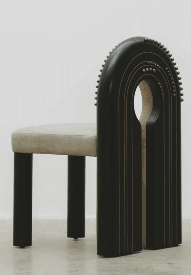 A contemporary chair with a distinctive U-shaped backrest that forms a cutout. The seat and the curved portion of the backrest are upholstered in a light beige, suede-like fabric. The frame is composed of dark, possibly black or dark wood, elements. The outer curved frame of the backrest and the vertical supports of the legs are adorned with a series of small, brass or gold-colored studs. The legs are thick and rectangular, with a dark finish. The chair's design is modern and somewhat sculptural, featuring a negative space in the upper backrest. — 3D model