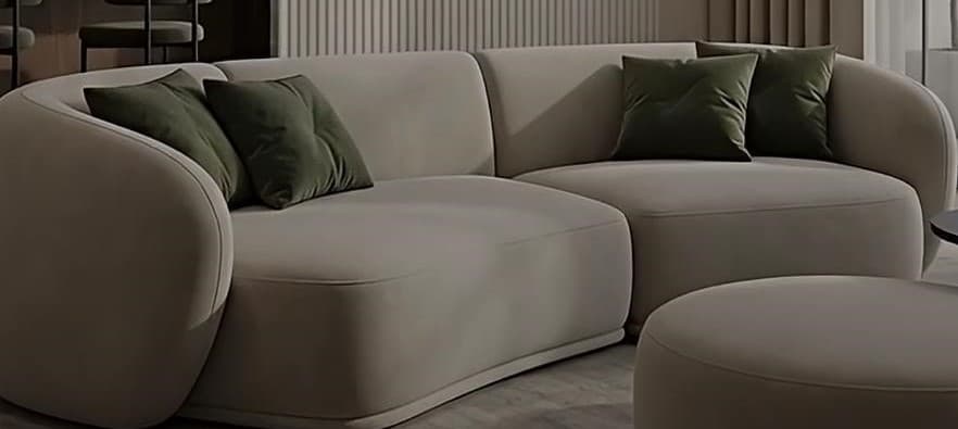 A modern, curved sectional sofa upholstered in a light beige fabric, accompanied by a matching round ottoman. It features soft, rounded lines and includes two dark green throw pillows. — 3D model