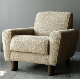 A modern upholstered armchair featuring a soft, textured bouclé fabric in an off-white or cream color. The chair has wide, rounded armrests and a slightly reclined backrest, contributing to a comfortable, inviting appearance. It is supported by four short, cylindrical dark wood legs. The overall design emphasizes softness and comfort with a contemporary aesthetic. — 3D model