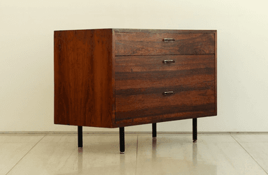 A dark wooden chest of three drawers with slender black legs and minimalist metal handles, set against a light wall. — 3D model