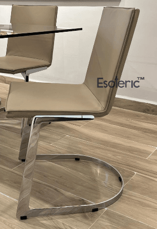 A modern dining chair with a sleek design. The seat and backrest are upholstered in a light beige material, likely leather or faux leather, with clean, squared-off edges. The frame is constructed from polished metal, featuring a cantilever base with a distinctive U-shaped support that extends from the legs and curves to form the base. The chair appears to be part of a set, with another similar chair visible in the background, partially obscured by a glass tabletop. — 3D model