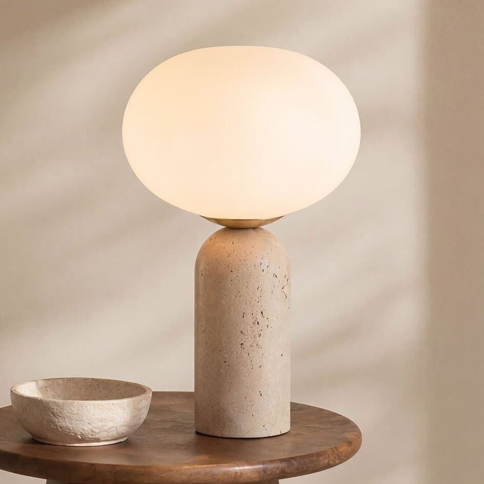 A modern table lamp featuring a cylindrical base made of textured, light-colored travertine, topped with a large spherical opaque white glass diffuser that emits a soft, ambient light. The design is minimalist and sculptural. — 3D model
