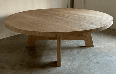 A round dining table with a thick, solid wood top and a substantial base constructed from heavy timber. The base features four angled legs connected by a cross-brace structure, providing stability and a rustic aesthetic. — 3D model