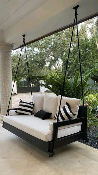 A modern suspended outdoor daybed featuring a rectangular black wooden frame, thick white upholstered cushions, and several throw pillows. The daybed is hung by four black metal rods or ropes from two overhead points, creating a swinging motion. The design is minimalist and elegant, suitable for a contemporary outdoor living space. — 3D model
